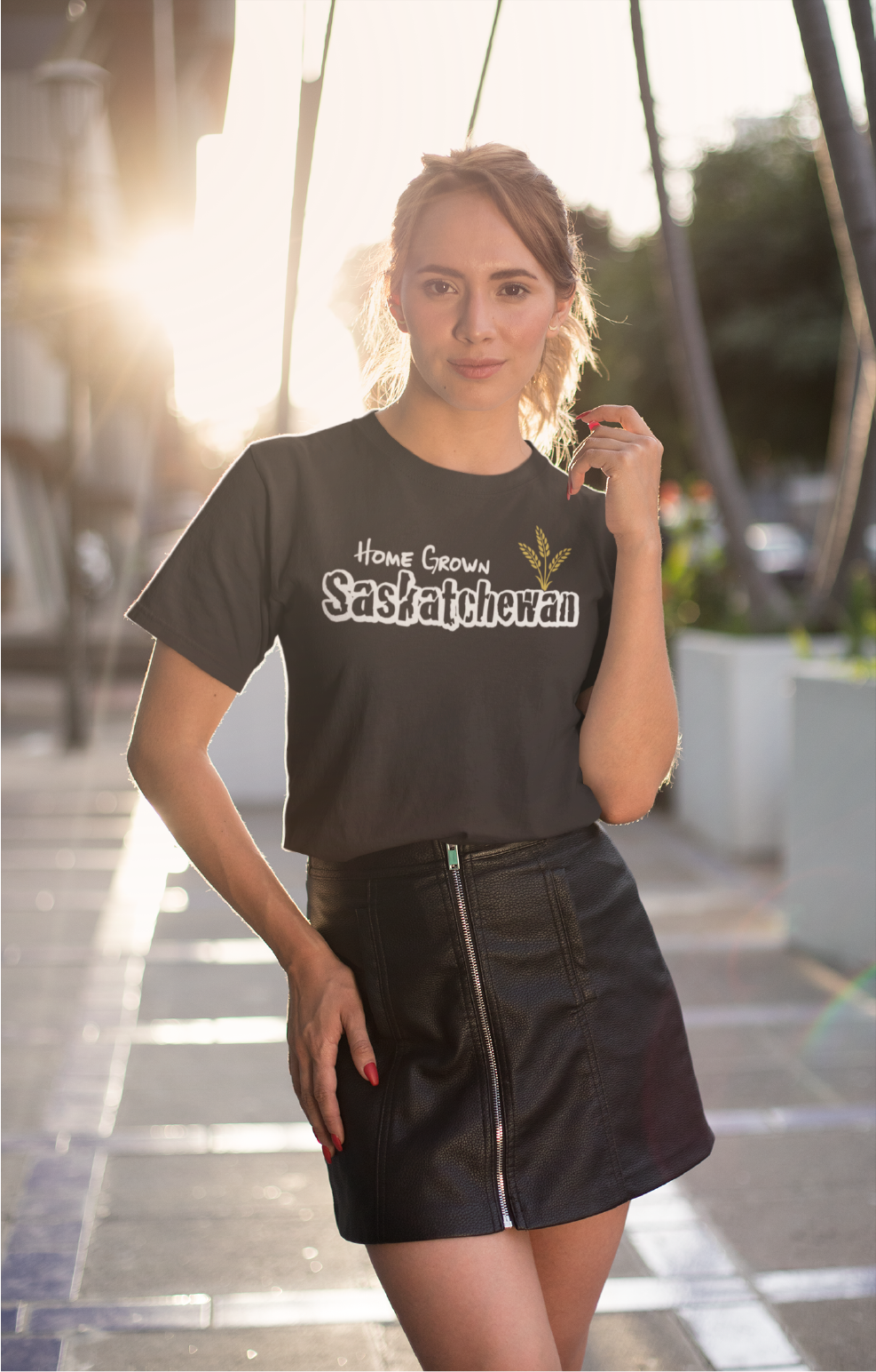 A young woman standing with the sunset behind her on a street wearing a skirt and a black t shirt with white writing that says home grown Saskatchewan with wheat on front chest