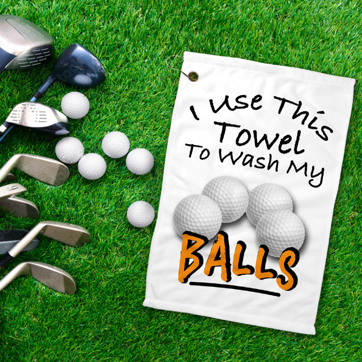 A 12x18 inch white golf towel laying on the grass next to golf clubs, that says in black writing Use this towel to wash my balls. With a picture of four golf balls 