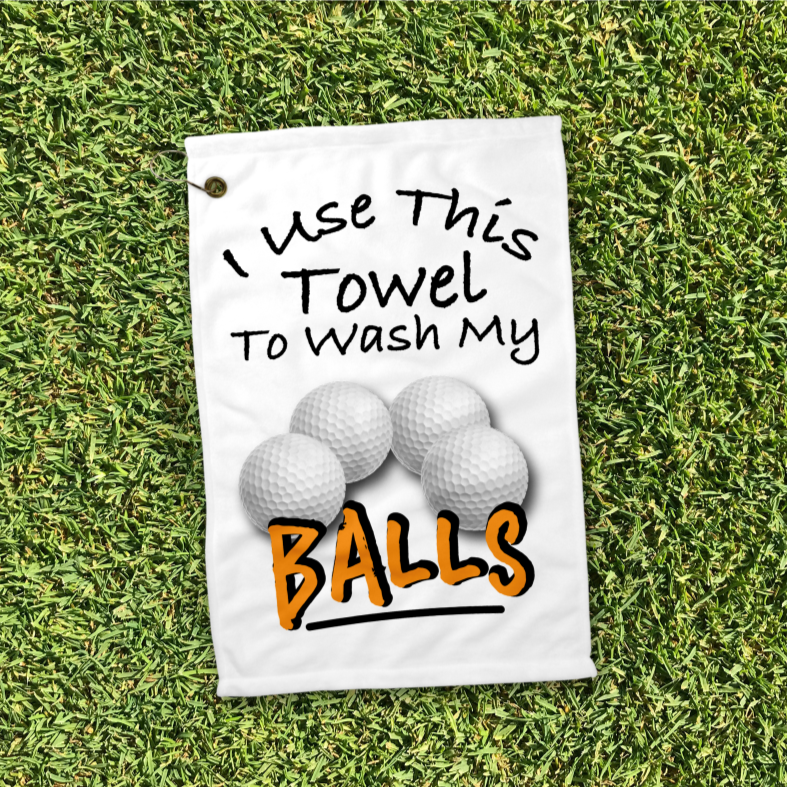 A 12x18 inch white golf towel laying on the grass that says in black writing Use this towel to wash my balls. With a picture of four golf balls 