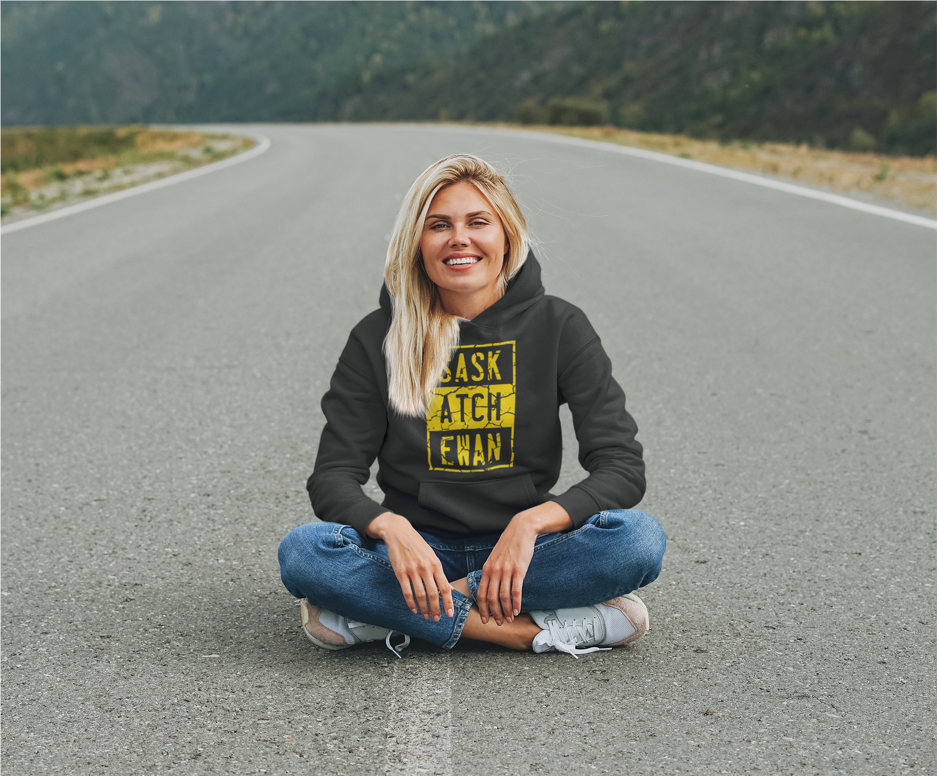 A young woman sitting in the middle of a highway wearing a black pullover hoodie with a yellow compton style Saskatchewan graphic on the front chest