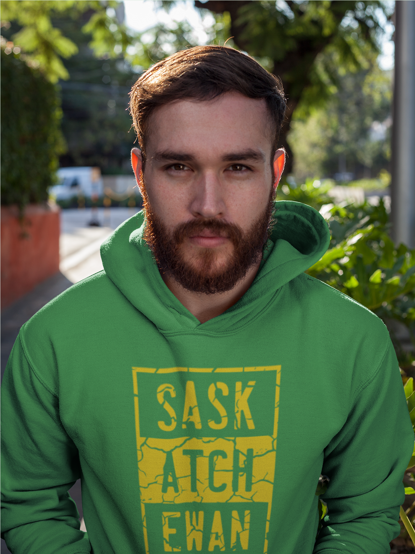 A man standing on a porch with his hands in side of the pouch of a green pullover hoodie with a yellow compton style Saskatchewan graphic on the front chest