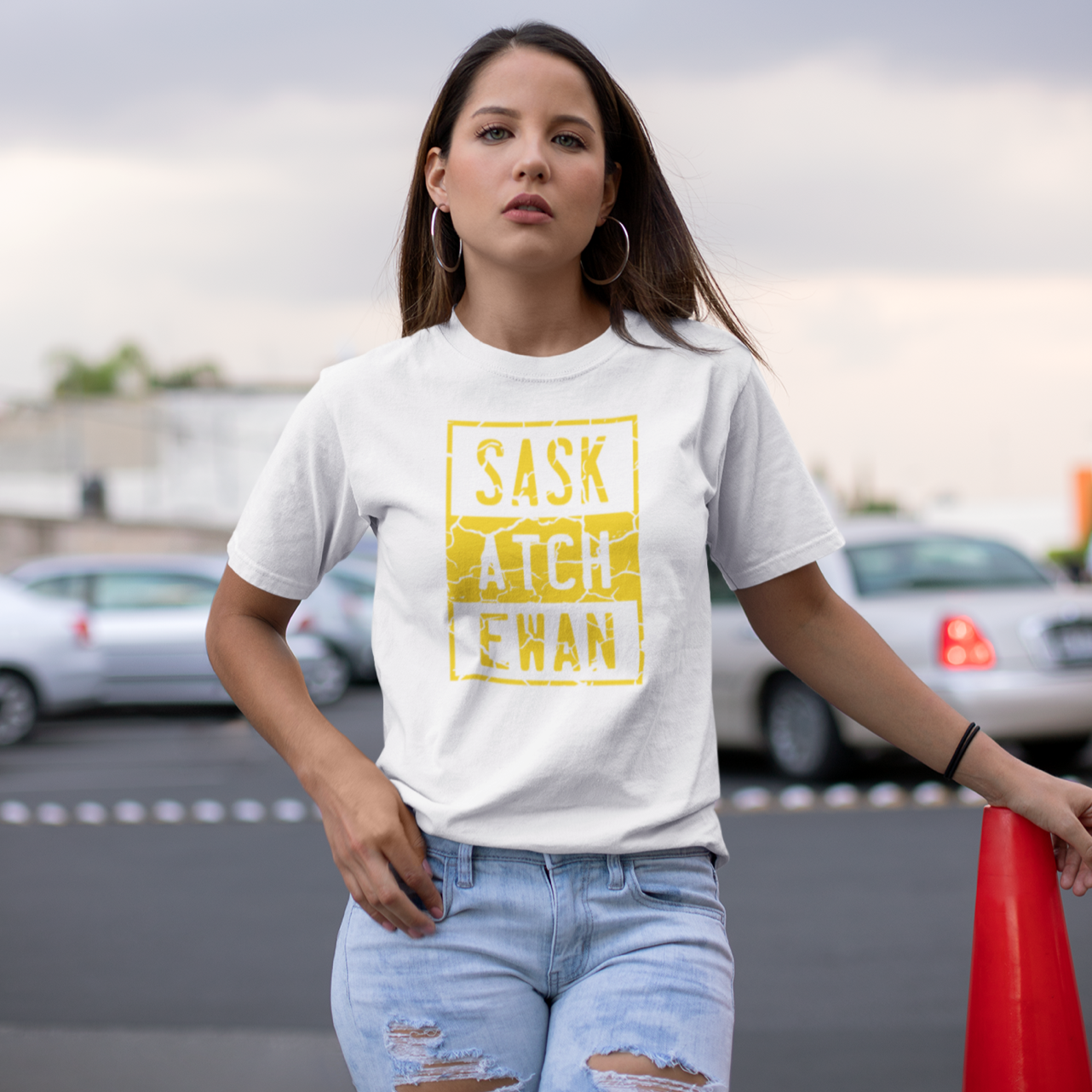 A young woman standing in a parking lot wearing torn jeans and a white t-shirt with a yellow compton style Saskatchewan graphic on the front chest