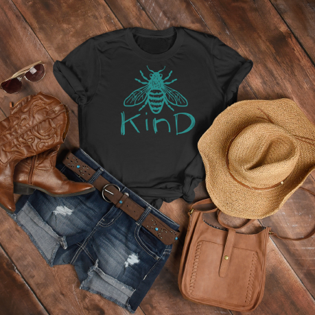 A women's black t shirt with a teal coloured bee and the word kind on the front chest, laying on a old wood floor surrounded by a cowgirl hat, boots and a purse.