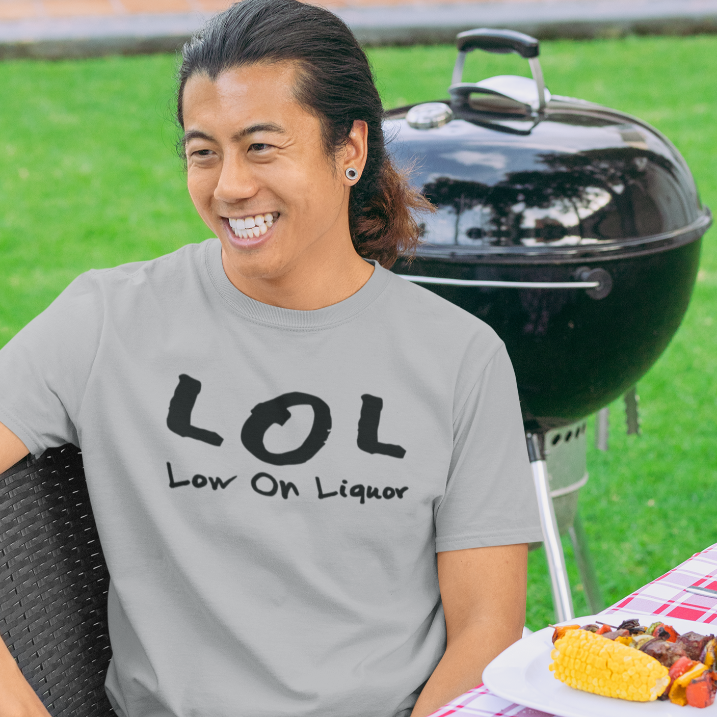 a smiling man by a grill at a bbq party wearing a grey t shirt with black phrase that say LOL low on liquor